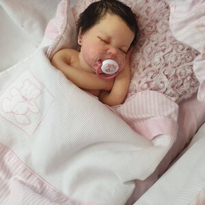 Pu&ograve; includere: Un bambolotto che dorme con capelli scuri e occhi marroni, avvolto in una coperta a righe rosa e bianche e con un ciuccio su cui &egrave; scritto "Milk".