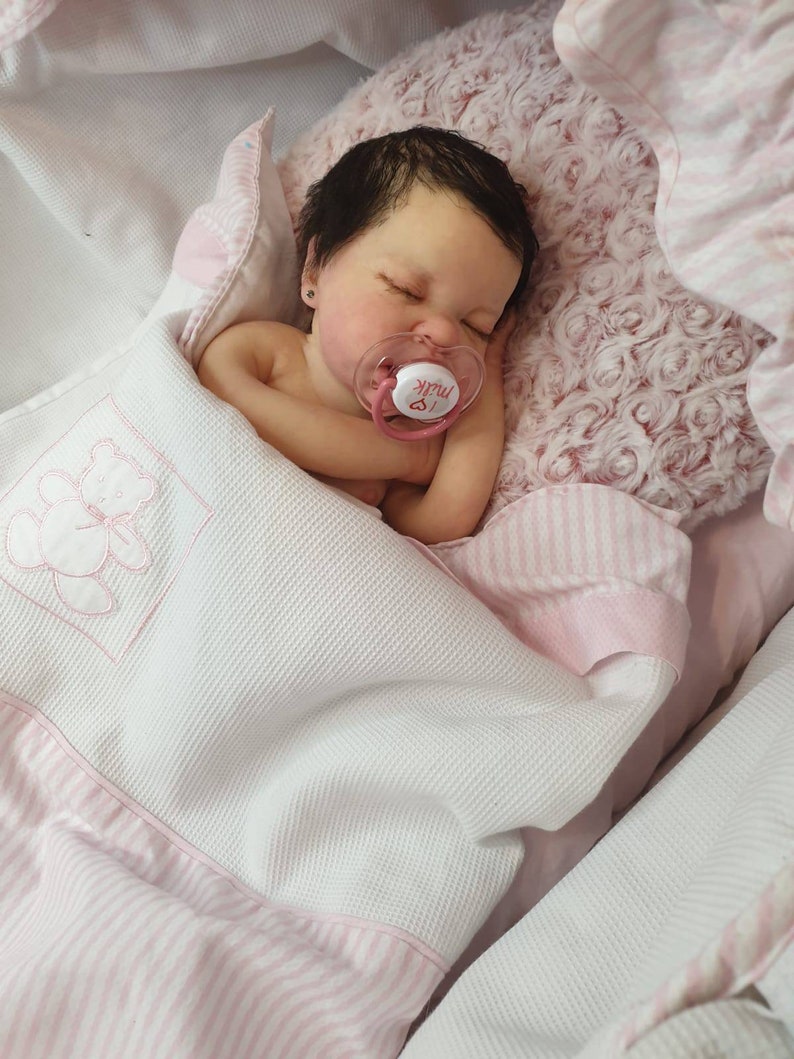 Pu&ograve; includere: Un bambolotto che dorme con capelli scuri e occhi marroni, avvolto in una coperta a righe rosa e bianche e con un ciuccio su cui &egrave; scritto "Milk".