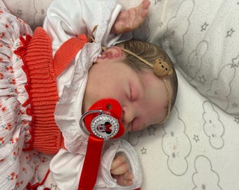 May include: A sleeping reborn baby doll wearing a white dress with a red knitted bodice and a red and white pacifier. The doll has a brown headband with a small brown bear charm.