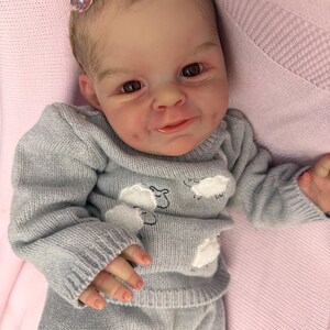 Puede incluir: Una mu&ntilde;eca beb&eacute; realista con un rostro detallado y ojos marrones. La mu&ntilde;eca viste un conjunto de jersey y pantal&oacute;n de punto gris, adornado con motivos de nubes blancas. Se ve un accesorio para el pelo rosa. La mu&ntilde;eca est&aacute; sobre una manta rosa.