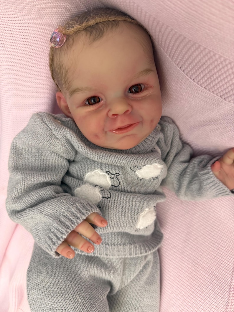 Puede incluir: Una mu&ntilde;eca beb&eacute; realista con un rostro detallado y ojos marrones. La mu&ntilde;eca viste un conjunto de jersey y pantal&oacute;n de punto gris, adornado con motivos de nubes blancas. Se ve un accesorio para el pelo rosa. La mu&ntilde;eca est&aacute; sobre una manta rosa.