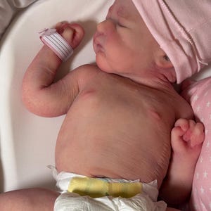 May include: A newborn baby wearing a pink beanie and a diaper with a yellow waistband. The baby has a hospital wristband and is lying in a white bassinet. The baby's skin is pink.