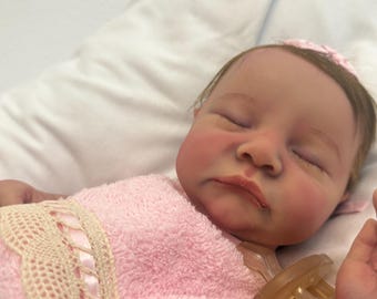 Puede incluir: Una muñeca bebé realista con los ojos cerrados, mejillas rosadas y un chupete. La muñeca está envuelta en una toalla rosa con detalles de encaje. Un lazo rosa adorna la cabeza de la muñeca. La muñeca está sobre una superficie blanca.