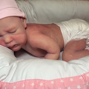 May include: A newborn baby doll wearing a pink knit cap and a diaper, lying on a white pillow with a pink star-patterned border. The doll's skin has a realistic appearance, with rosy cheeks and closed eyes.