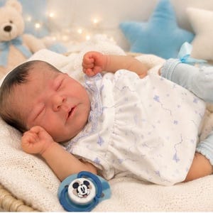 May include: A realistic newborn baby doll with dark hair, sleeping with eyes closed. The doll wears a white sleeveless dress with a small blue kite pattern, light blue ruffled socks with ribbons, and lies next to a blue and white Mickey Mouse pacifier.
