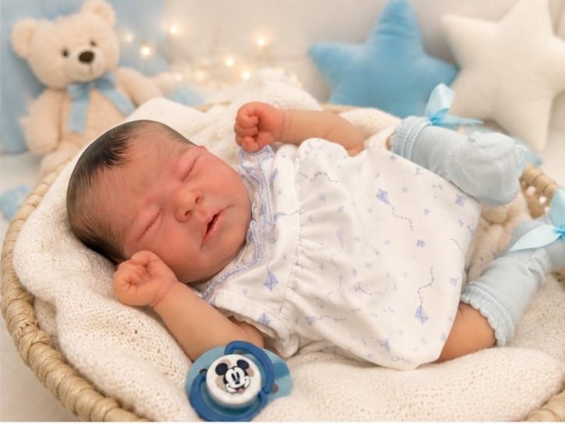 May include: A realistic newborn baby doll with dark hair, sleeping with eyes closed. The doll wears a white sleeveless dress with a small blue kite pattern, light blue ruffled socks with ribbons, and lies next to a blue and white Mickey Mouse pacifier.