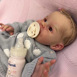 Puede incluir: Una mu&ntilde;eca beb&eacute; realista con un su&eacute;ter gris con detalles blancos y un chupete. Una botella para beb&eacute;s con l&iacute;quido rosa est&aacute; al lado de la mu&ntilde;eca. La mu&ntilde;eca est&aacute; acostada sobre una manta rosa.