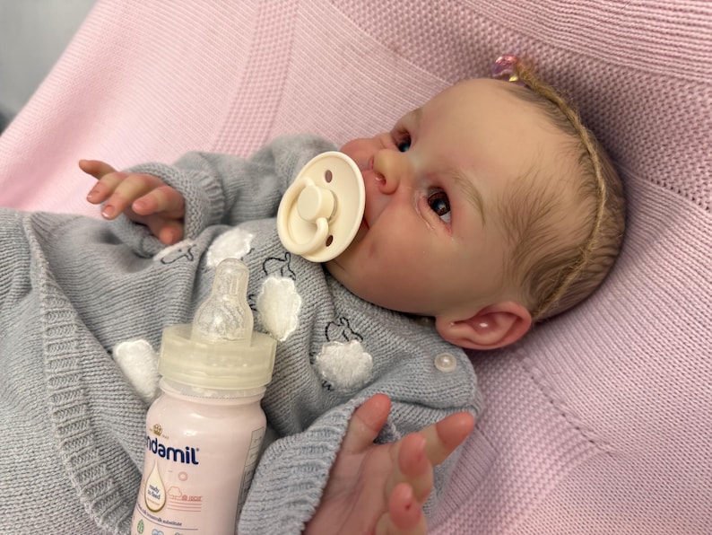 Puede incluir: Una mu&ntilde;eca beb&eacute; realista con un su&eacute;ter gris con detalles blancos y un chupete. Una botella para beb&eacute;s con l&iacute;quido rosa est&aacute; al lado de la mu&ntilde;eca. La mu&ntilde;eca est&aacute; acostada sobre una manta rosa.