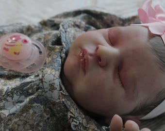 May include: A sleeping baby doll with a pink bow and a pacifier. The doll is wearing a floral dress and has realistic features.