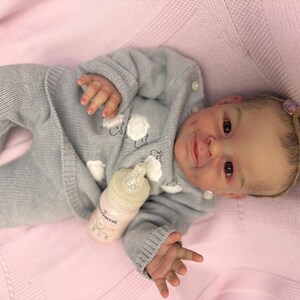 Puede incluir: Una mu&ntilde;eca beb&eacute; realista con un conjunto de punto gris con aplicaciones de ovejas blancas, acostada sobre una manta rosa. La mu&ntilde;eca tiene un biber&oacute;n y un peque&ntilde;o accesorio rosa para el pelo. La piel de la mu&ntilde;eca tiene un tono realista.