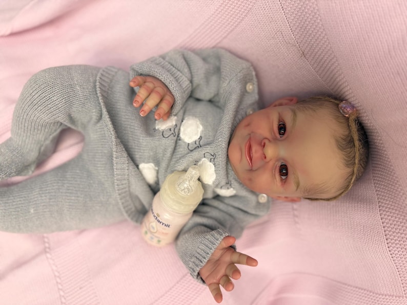 Puede incluir: Una mu&ntilde;eca beb&eacute; realista con un conjunto de punto gris con aplicaciones de ovejas blancas, acostada sobre una manta rosa. La mu&ntilde;eca tiene un biber&oacute;n y un peque&ntilde;o accesorio rosa para el pelo. La piel de la mu&ntilde;eca tiene un tono realista.