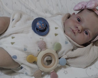 May include: A baby doll with blue eyes and a pink bow headband is lying on a white blanket with a pacifier and a teether toy.