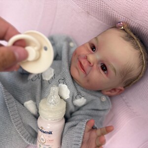 Puede incluir: Una mu&ntilde;eca beb&eacute; realista con un su&eacute;ter gris con detalles de nubes blancas, acostada sobre una manta rosa. Se sostiene un chupete cerca de la cara de la mu&ntilde;eca y se ve un biber&oacute;n de f&oacute;rmula. La mu&ntilde;eca tiene una diadema con un lazo rosa.