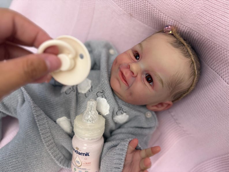 Puede incluir: Una mu&ntilde;eca beb&eacute; realista con un su&eacute;ter gris con detalles de nubes blancas, acostada sobre una manta rosa. Se sostiene un chupete cerca de la cara de la mu&ntilde;eca y se ve un biber&oacute;n de f&oacute;rmula. La mu&ntilde;eca tiene una diadema con un lazo rosa.