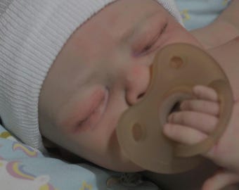 May include: A sleeping baby doll wearing a white knitted hat and a brown dummy.