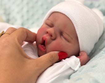 Puede incluir: Una muñeca bebé realista con gorro de punto blanco y body blanco. La muñeca tiene mejillas rosadas y ojos cerrados. Una mano con un anillo dorado y esmalte de uñas rojo toca suavemente la cara de la muñeca.