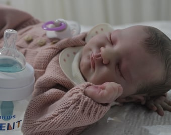May include: A pink knitted baby doll with closed eyes, wearing a white bib with pink polka dots. A clear baby bottle with a blue liquid inside is in the foreground.