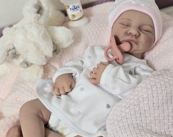 May include: A sleeping baby doll wearing a pink and white hat, a white onesie with pink hearts, and a pacifier. The doll is lying on a pink blanket with a white stuffed animal nearby.