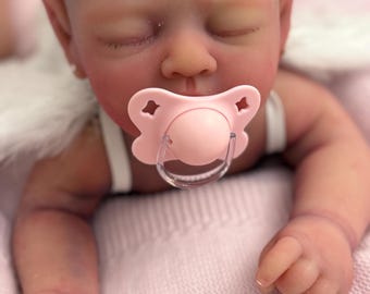 Puede incluir: Una muñeca bebé realista con los ojos cerrados y un chupete rosa. La muñeca lleva una diadema floral y está tumbada sobre una manta rosa. La piel de la muñeca tiene un tono realista.