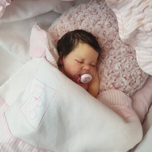 Pu&ograve; includere: Un bambolotto che dorme con capelli scuri e occhi marroni &egrave; sdraiato su una coperta a righe rosa e bianche. Il bambolotto indossa un ciuccio rosa con la scritta "Milk".