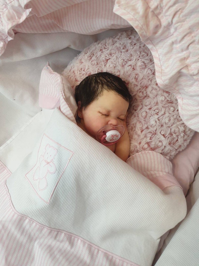Pu&ograve; includere: Un bambolotto che dorme con capelli scuri e occhi marroni &egrave; sdraiato su una coperta a righe rosa e bianche. Il bambolotto indossa un ciuccio rosa con la scritta "Milk".