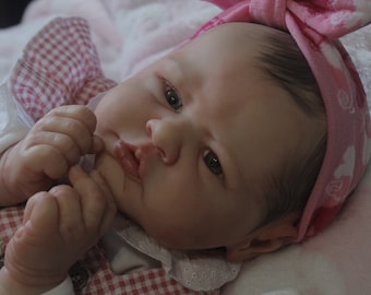 May include: A close-up of a baby doll with brown hair wearing a pink and white headband and a pink and white checkered dress.