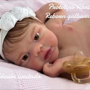 Może przedstawiać: Realistyczna lalka dla dziecka z brązowymi oczami i białą opaską z kwiatami. Lalka leży na różowym kocu i ma smoczek w buzi. Widoczny jest tekst "Prototipo Abril Reborn galbanno" i "Edición limitada".