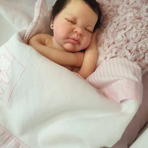 Pu&ograve; includere: Un bambolotto che dorme con i capelli scuri e gli occhi chiusi, sdraiato su una coperta a righe rosa e bianche con una coperta bianca sopra. Il bambolotto indossa un body bianco.