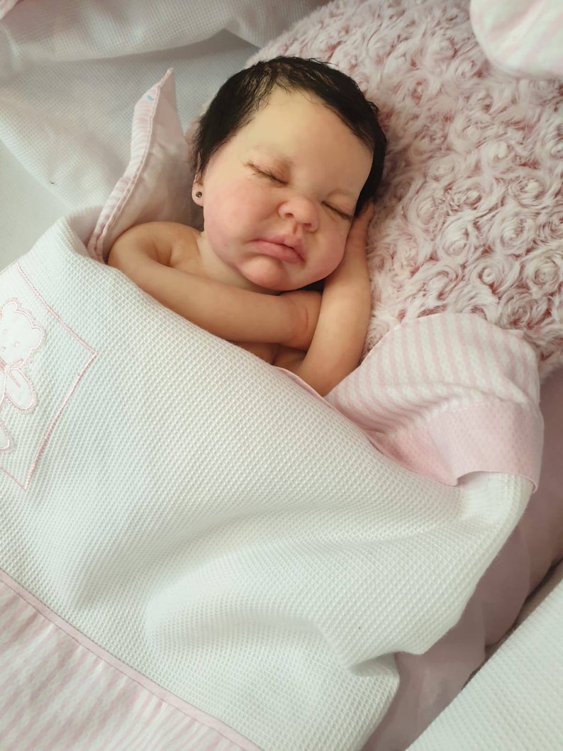 Pu&ograve; includere: Un bambolotto che dorme con i capelli scuri e gli occhi chiusi, sdraiato su una coperta a righe rosa e bianche con una coperta bianca sopra. Il bambolotto indossa un body bianco.