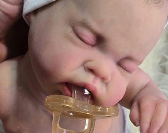 May include: A close-up of a baby doll with its eyes closed, wearing a white headband and a pacifier in its mouth.