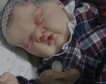 May include: A sleeping baby doll wearing a blue and red plaid dress with a white lace collar and a white headband with silver accents. The doll is holding a pacifier.