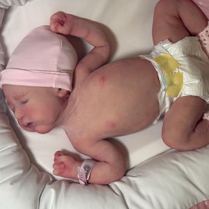 May include: A newborn baby wearing a pink cap, diaper, and pink crocheted booties, lying on a white surface. The baby has a hospital wristband. The baby is lying on a white pillow.