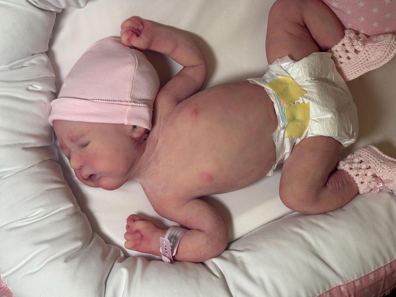 May include: A newborn baby wearing a pink cap, diaper, and pink crocheted booties, lying on a white surface. The baby has a hospital wristband. The baby is lying on a white pillow.