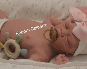 Puede incluir: Una muñeca reborn con un lazo rosa en la cabeza, que lleva un gorro blanco y un pañal. La muñeca está tumbada sobre una manta blanca con un patrón de estrellas. La muñeca está sujetando un chupete en la boca y tiene un juguete en la mano. El texto "Reborn Galbarro" es visible en el pecho de la muñeca.
