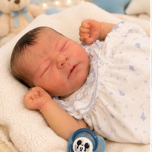 May include: A realistic newborn doll with dark hair, sleeping with eyes closed. The doll wears a white short-sleeved dress featuring a light blue kite pattern and a ruffled collar. A blue pacifier with a Mickey Mouse graphic rests near the doll.