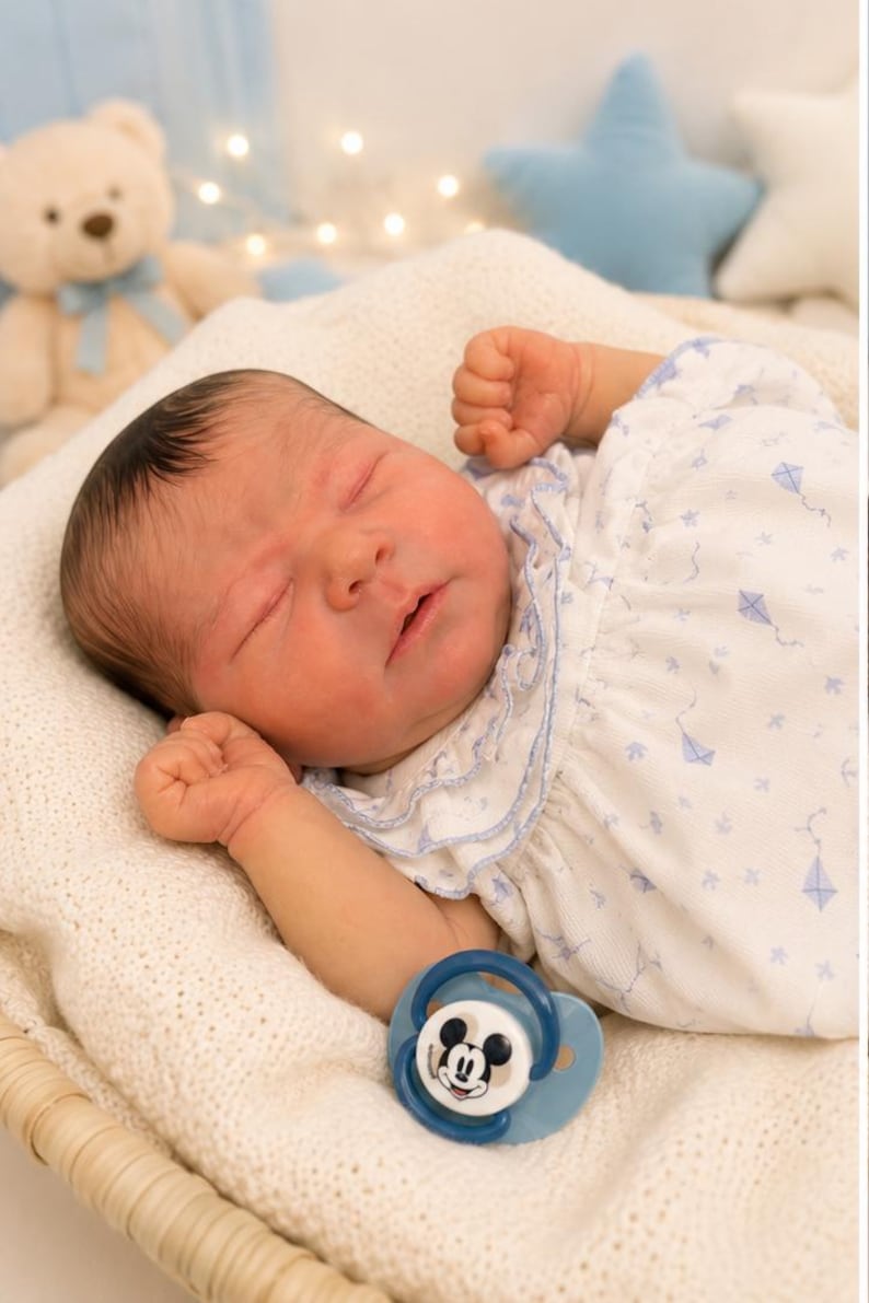 May include: A realistic newborn doll with dark hair, sleeping with eyes closed. The doll wears a white short-sleeved dress featuring a light blue kite pattern and a ruffled collar. A blue pacifier with a Mickey Mouse graphic rests near the doll.