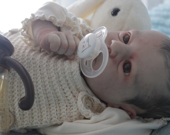 May include: A realistic baby doll with brown hair, wearing a white knitted sweater and a pacifier. The doll is lying on a blue and white blanket.
