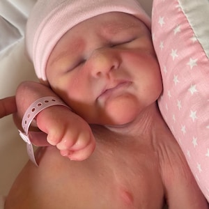 May include: A newborn baby wearing a pink beanie and a hospital wristband, resting on a white surface. The baby's face is flushed, and it is next to a pink pillow with white star patterns.