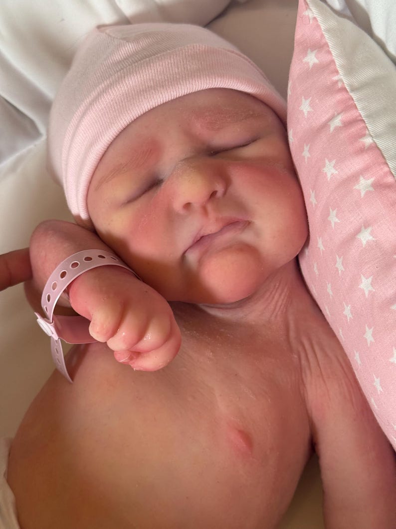May include: A newborn baby wearing a pink beanie and a hospital wristband, resting on a white surface. The baby's face is flushed, and it is next to a pink pillow with white star patterns.