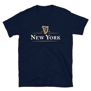 May include: A navy blue t-shirt featuring a gold harp logo and the text "NEW YORK" underneath. The harp logo has the numbers "17" and "59" on either side.