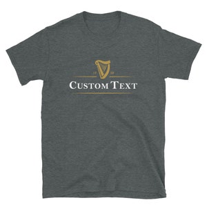 Op de afbeelding: Donkergrijs t-shirt met een gouden harp logo en de jaartallen 1759 en 1759. De tekst "CUSTOM TEXT" is in het wit onder het logo gedrukt. Het shirt is gemaakt van een zacht materiaal.