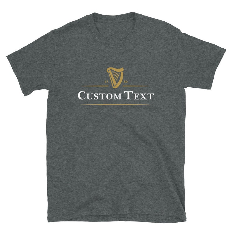 Op de afbeelding: Donkergrijs t-shirt met een gouden harp logo en de jaartallen 1759 en 1759. De tekst "CUSTOM TEXT" is in het wit onder het logo gedrukt. Het shirt is gemaakt van een zacht materiaal.