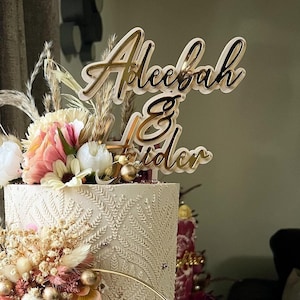 May include: A white wedding cake decorated with dried flowers and greenery. The cake is topped with a wooden sign that says "Aleezah & Tyler".