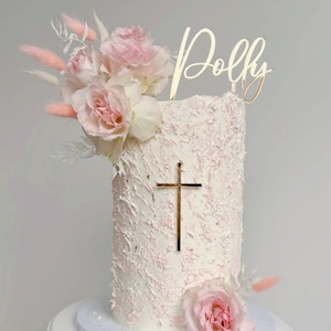 May include: A white cake with pink frosting and a gold cross topper. The cake is decorated with pink flowers and a wooden topper that says "Polly".