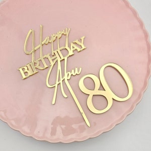 Pode incluir: Topo de bolo em acrílico dourado com as palavras "Feliz aniversário" e o número "80".