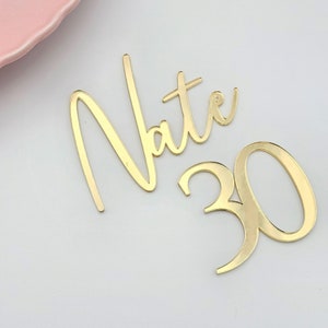 Pode incluir: Topo de bolo de acrílico dourado espelhado com nome e número. O nome "Nate" está escrito em letra cursiva e o número "30" está em negrito.