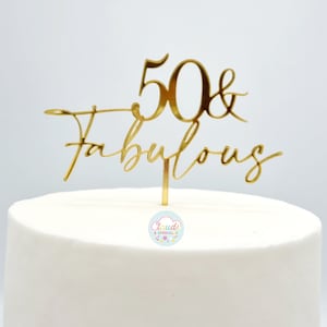 以下が含まれることがあります： 「50 & Fabulous」というテキストが書かれた、筆記体フォントの金のケーキトッパー。トッパーは白いケーキの上に置かれています。