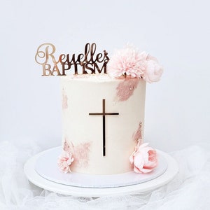 Mirror gold acrylic name topper with a spike stuck into the top of a cake. The topper has the name in a script font with "BAPTISM" written in a block font underneath. There is a mirror gold acrylic crucifix cake charm stuck on the front of the cake.