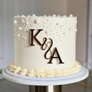 Puede incluir: Una tarta glaseada blanca adornada con perlas comestibles y un monograma "K & A" dorado. La tarta está sobre un soporte blanco, con un borde decorativo de glaseado color crema.
