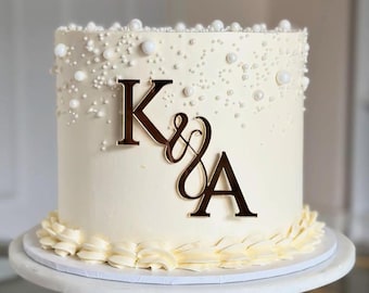 Puede incluir: Una tarta glaseada blanca adornada con perlas comestibles y un monograma "K & A" dorado. La tarta está sobre un soporte blanco, con un borde decorativo de glaseado color crema.